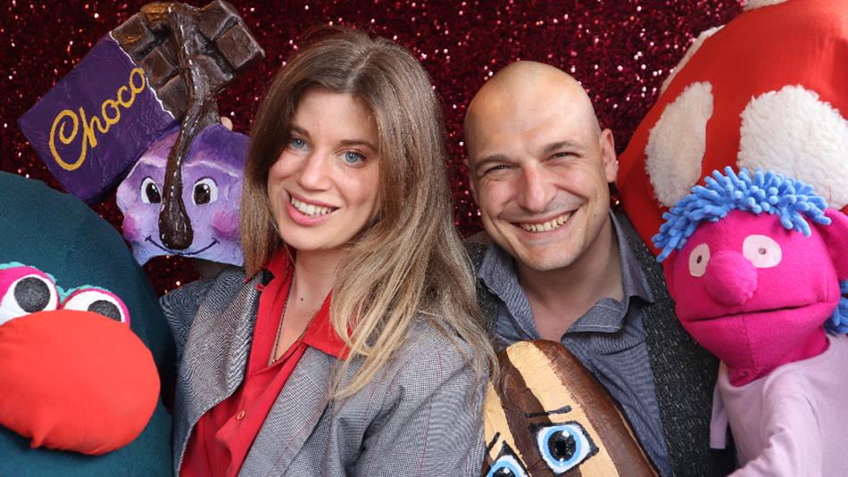 Two performers smiling at the camera surrounded by colourful puppets.