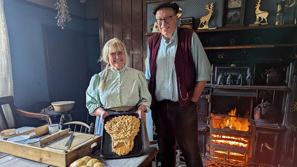 Two people stand in a recreated pit cottage, in front of a coal-fired range. One holds a bread display for Harvest Celebration.