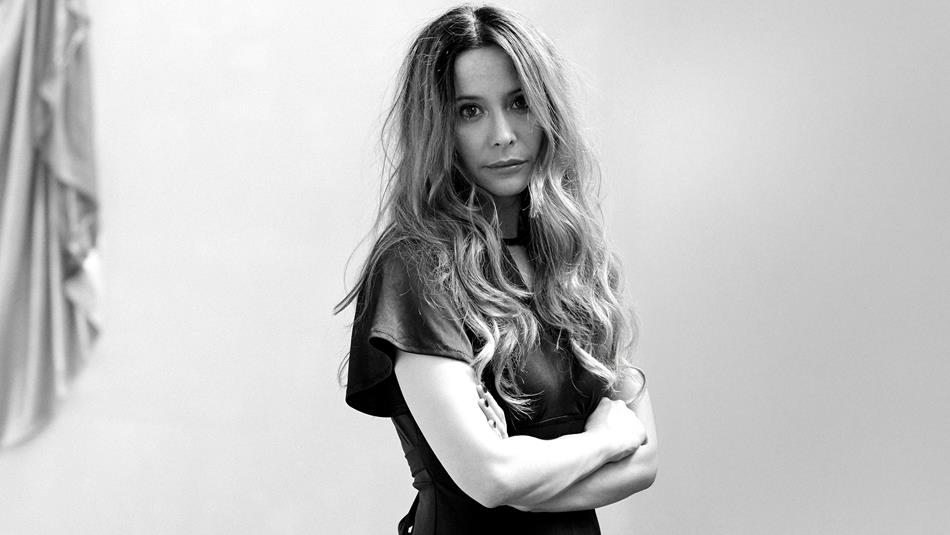 Black and white photograph of a woman with long wavy hair standing with arms crossed, facing slightly to the side. Background is plain with a draped f