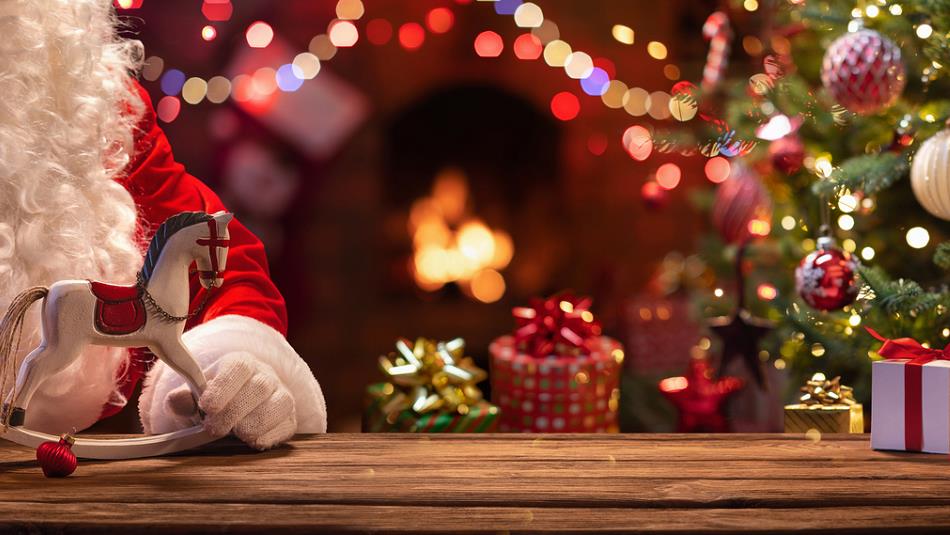 Santa holding a miniature rocking horse, and a Christmas tree, fairy lights and presents in the background.