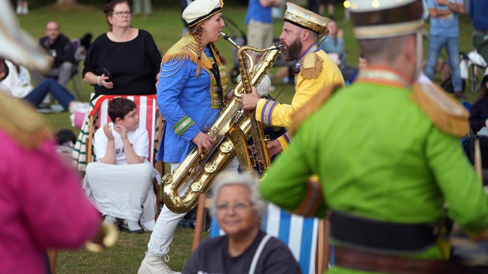 Brass musicians performing to crowds outdoors and people sitting and enjoying the show on deck chairs around them.