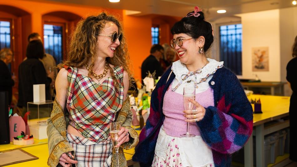 Photograph of two women socialising indoors at a brightly lit event or gathering, each holding a glass of champagne. The setting features yellow table