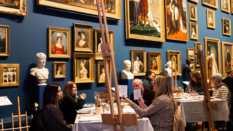 Photograph of an art gallery room with people seated at tables engaged in painting or drawing activities. Walls are adorned with framed artworks.