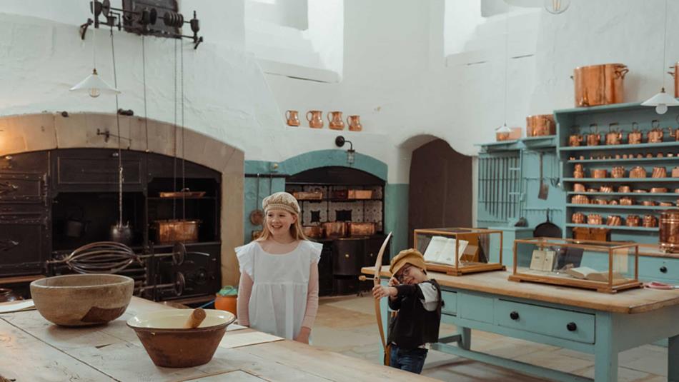 Two children dressed as servants in the kitchen at Raby Castle. The little girl stands next to a mixing bowl, and the boy has a bow and arrow.
