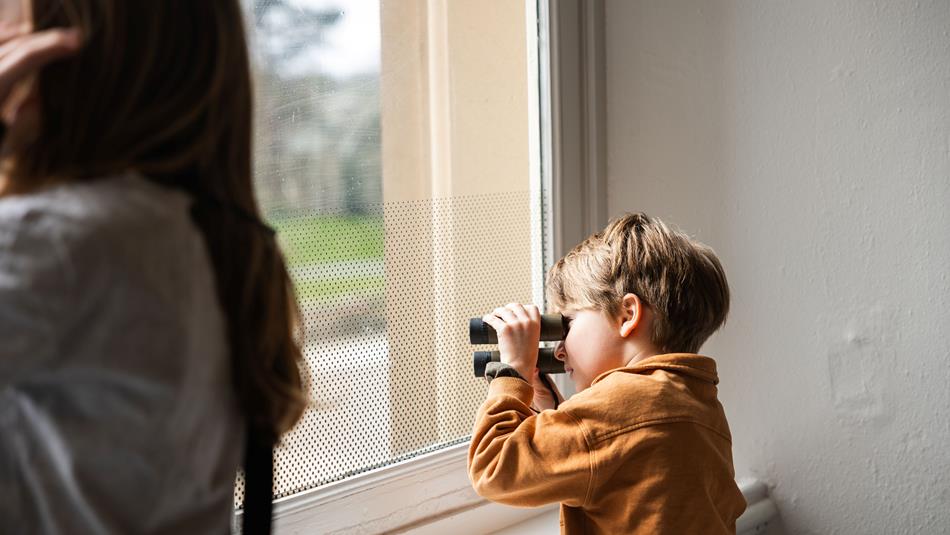 A young boy looking through binoculars out of a window, with a blurred figure of a woman in the foreground.