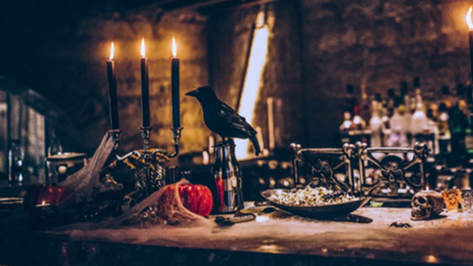 A raven sits at a dinner table covered in cobwebs, candles, skulls and pumpkins.