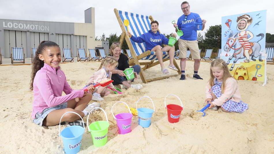 Adults and children enjoying the pop-up beach at Locomotion by Richard Salkeld, with colourful buckets and spades, a giant deckchair and seaside photo