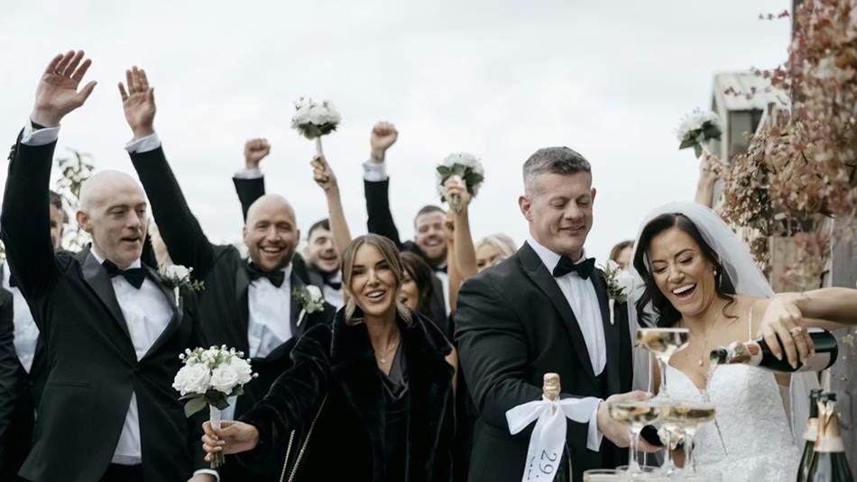 Bride and Groom smiling and pouring drinks, and their wedding party celebrating with their hands in the air behind them. Photo by Lauren Stacey.