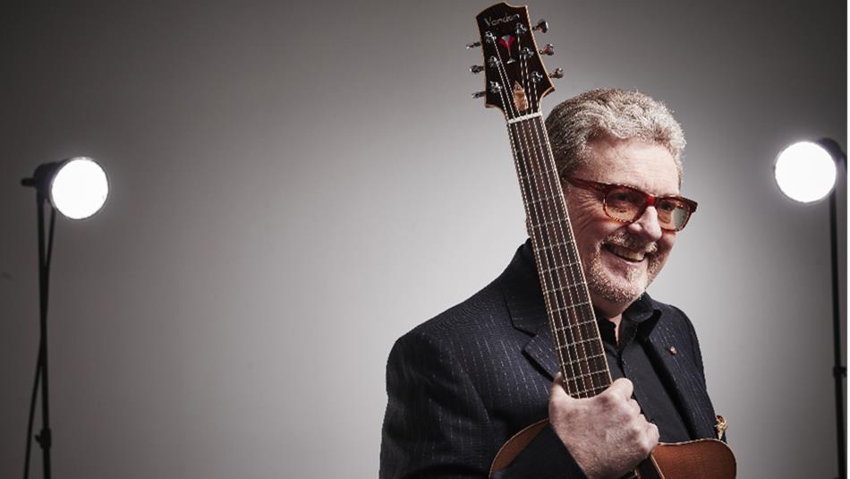 A man holding an acoustic guitar vertically against his shoulder, set in a studio with two bright lights positioned behind him on either