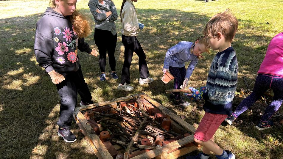 A group of children gathered around a wooden frame filled with sticks and bricks on grass, engaged in a hands-on outdoor activity.