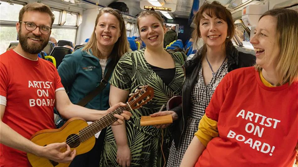 Sam Slatcher smiling and playing guitar on public transport, standing with four women who are also smiling.