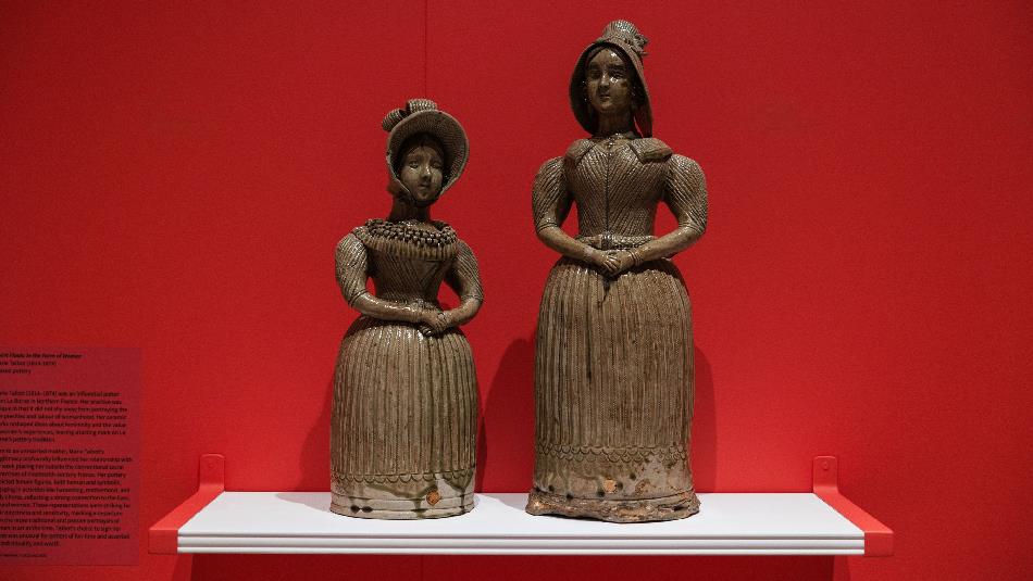 Two ceramic spirit flasks shaped like women in 19th-century attire stand on a white shelf against a vivid red wall, showcasing intricate detailing in