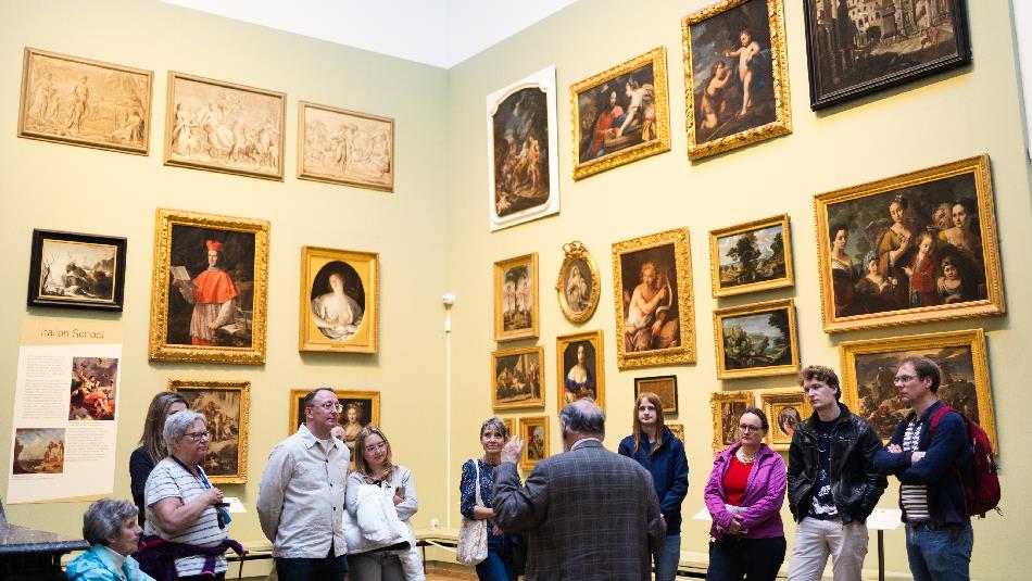 A group of ten people listening attentively to a curator in a museum gallery filled with framed historical paintings.
