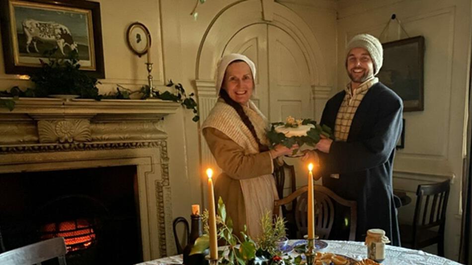 Two members of staff in Georgian clothing, holding Christmas cake.
