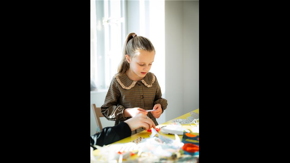 A young girl in a brown dress with hair in a ponytail, holding something made of paper, with craft materials on a yellow table in front