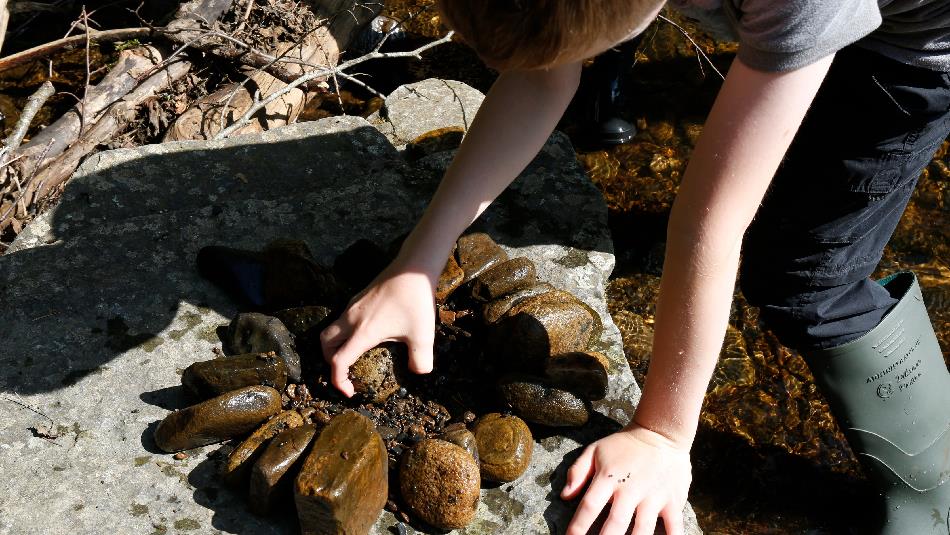 A child in rubber boots leans over a rock by a clear stream, arranging small stones into a circular pattern