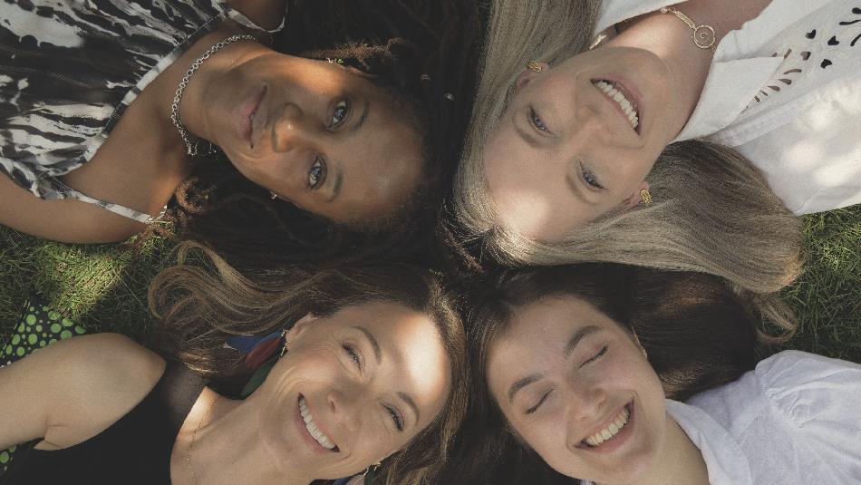 Four women lying on grass in a circle with heads close together, viewed from above. They wear casual clothing in black, white, and patterned fabrics.