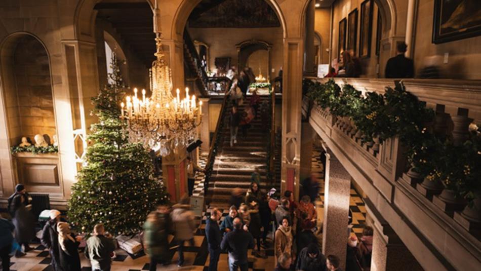 Photograph of a grand interior space with festive decoration, featuring a large Christmas tree with lights and ornaments, a chandelier, and garlands.