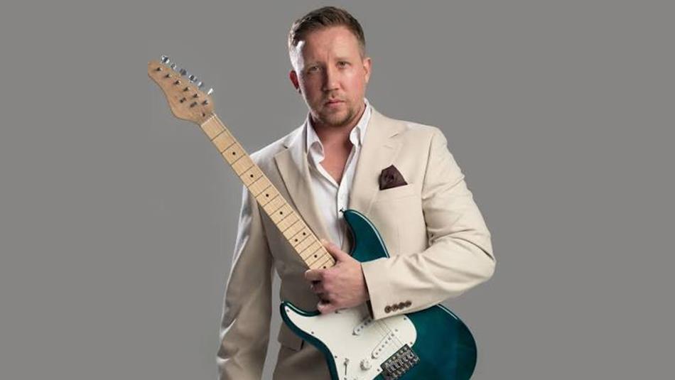 Luke Palmer, wearing a cream suit, holding a teal electric guitar.