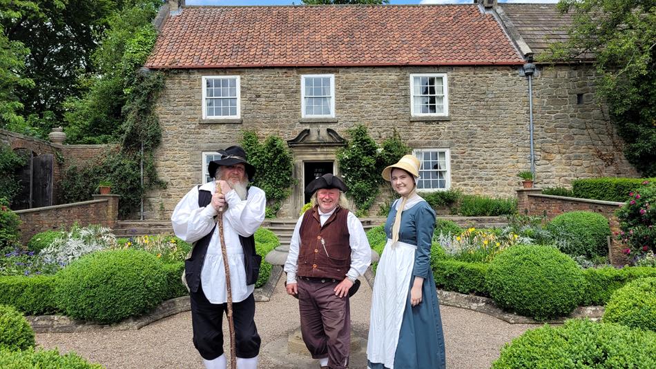 Three people in Georgian costume stand in front of Pockerley Old Hall at Beamish Museum.
