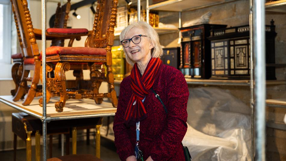 A smiling woman in a red outfit stands in a museum storeroom surrounded by intricately carved antique furniture and ornate wooden cabinets