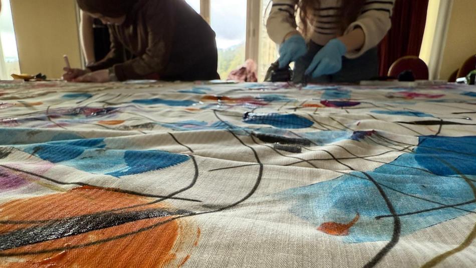Two people working on a large fabric with colourful abstract patterns in blue, orange, and black lines. Focus is on fabric surface.