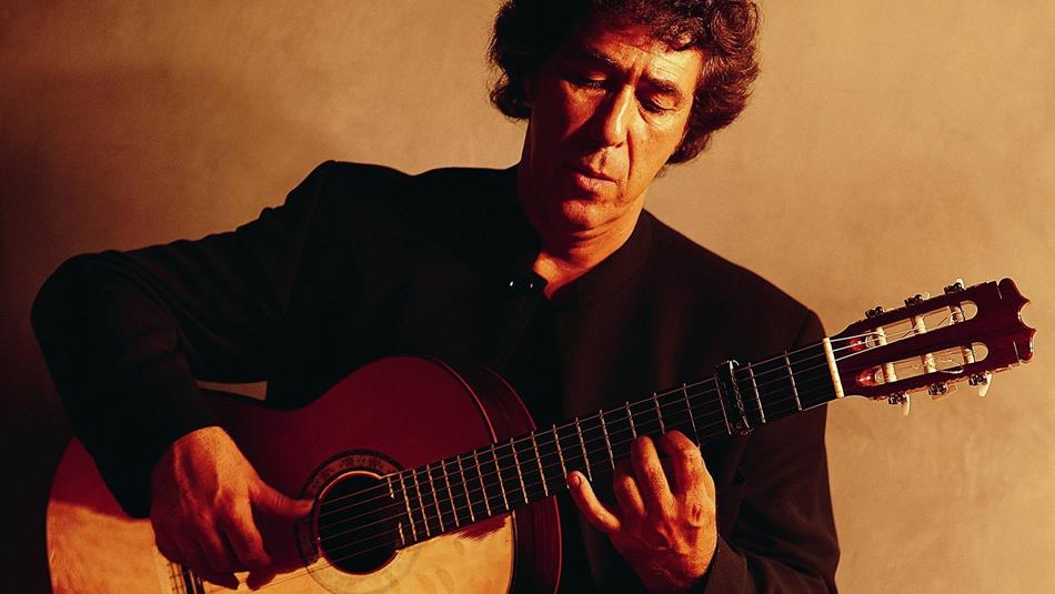 A man playing a classical guitar, seated against a plain, warm-toned background. The man wears a dark suit, focusing on his fingers.