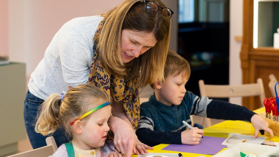A woman leans over a table helping two young children with an arts-and-crafts activity. The children sit at a bright yellow table covered with paper,