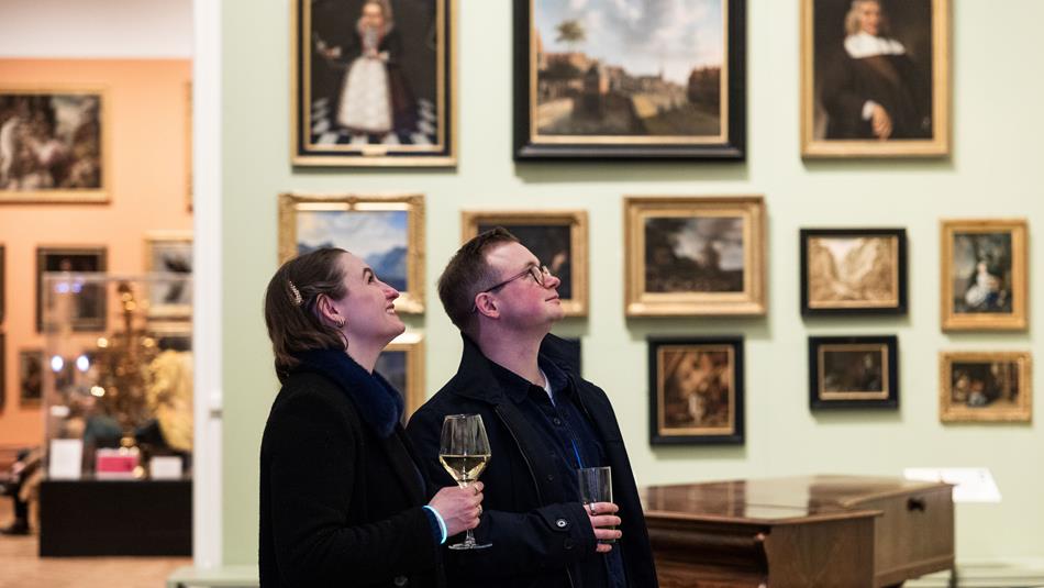 Photograph of two people holding drinks and observing framed paintings displayed on a gallery wall. The setting includes a grand piano and art.