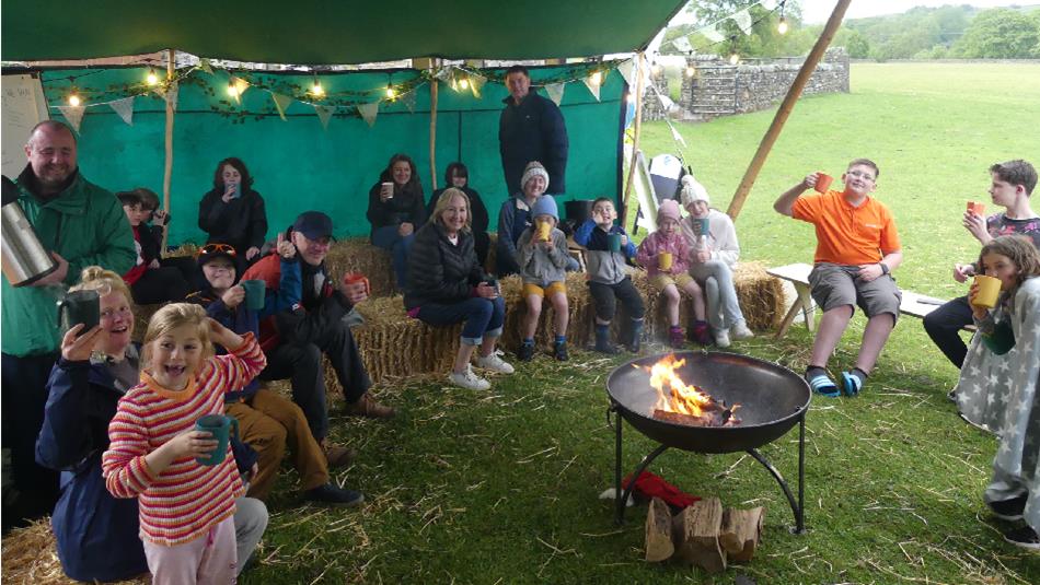 Families and children relax on hay bales under a green canopy tent, holding cups of hot drinks and gathered around a small fire pit, with open country