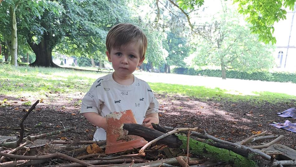 Child sitting in the grounds of The Bowes Museum , holding a plant pot,  surrounded by twigs and trees.