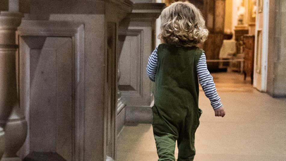 A young child with curly blonde hair, wearing green dungarees and a striped shirt, walks alone along a grand, ornately decorated museum hallway