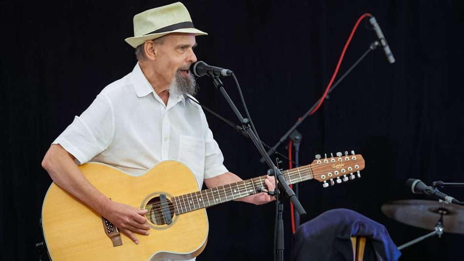Mark Harrison wearing a hat and white shirt, performing on stage with his guitar.