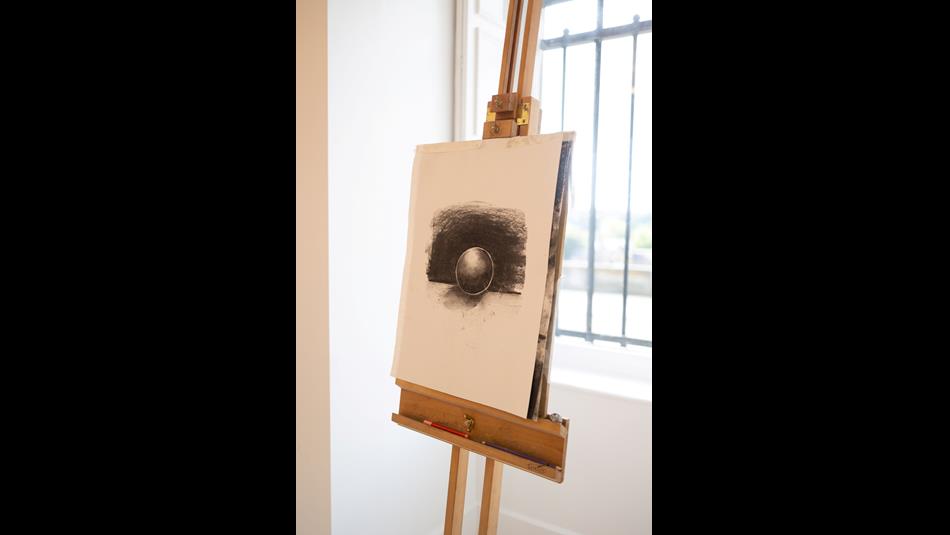 A wooden easel holding a drawing pad with a charcoal sketch of a sphere against a dark shaded background. Natural light from a window behind.