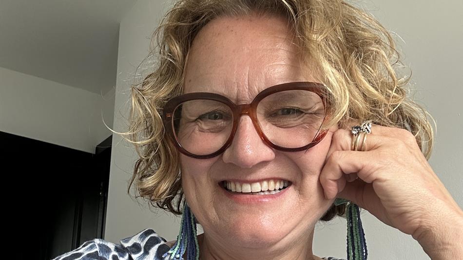 Smiling person with short, curly blonde hair and round brown glasses, resting their chin on one hand. They are wearing a patterned blue-and-white top