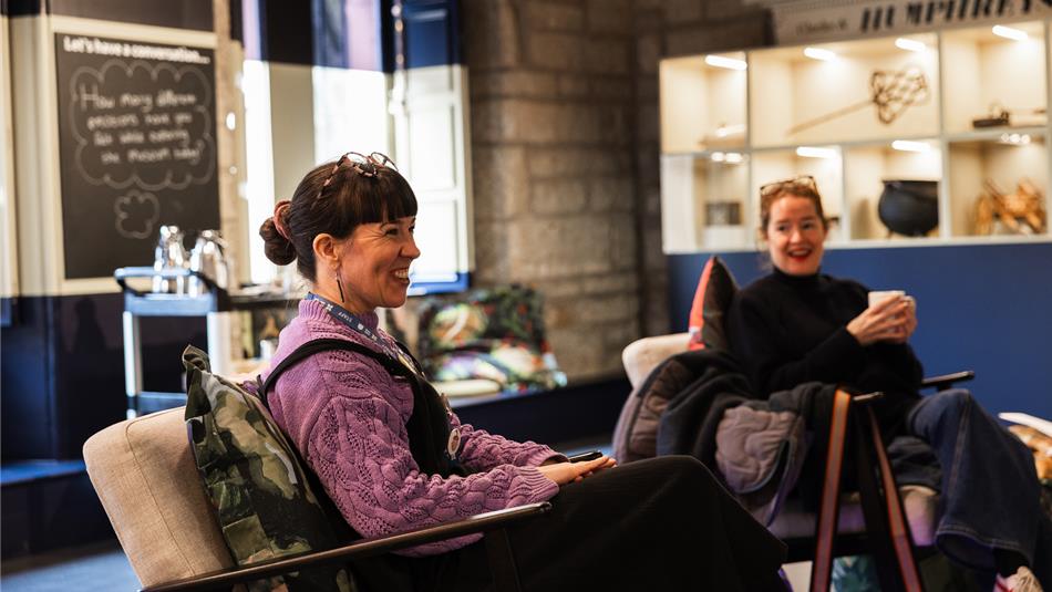 Two people seated indoors in a cozy, well-lit room with stone and blue walls, and large windows. One person wears a purple sweater and s