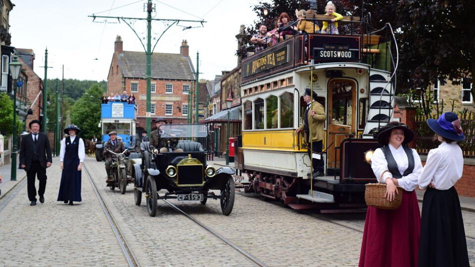 Staff in character at Beamish Museum, next to vintage cars and a tram - 1900s Town