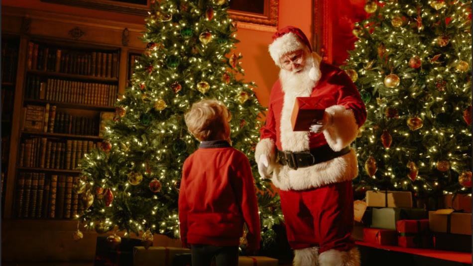 Santa giving a present to a small child in front of two ornate Christmas trees