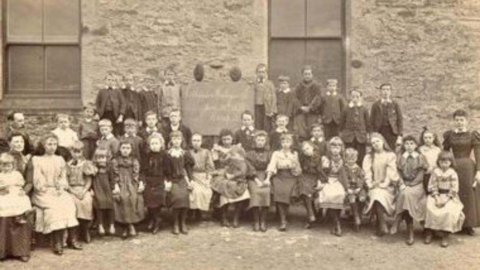 An old fashioned school photograph of children sat in lines in front of a school building
