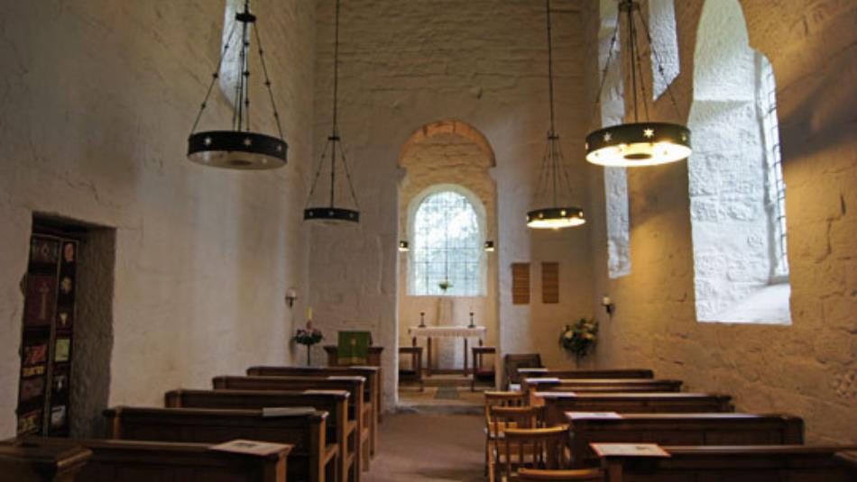 The interior of an Anglo Saxon church, with white stone walls and hanging round lights.