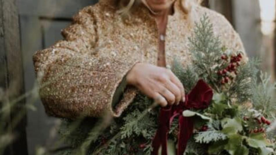 A person, shoulders down, making a Christmas wreath with a burgundy bow