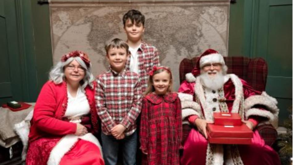 3 children standing whilst Mr and Mrs Clause sit beside them with red presents