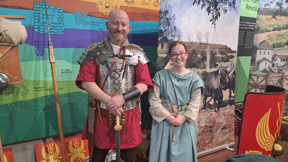 Two people dressed in Roman clothes, smiling in front of information boards