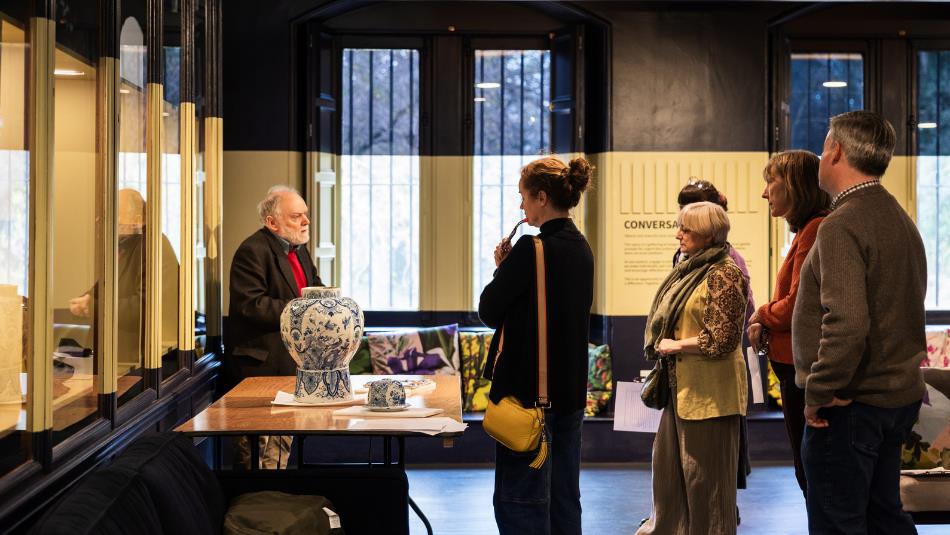 Photograph of a museum or gallery setting where a guide explains exhibits to a group of four visitors. The scene features display cases with artifacts