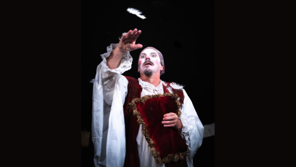 A person on a dark stage wearing dramatic white face pain in historical robes, reaching out for a feather.