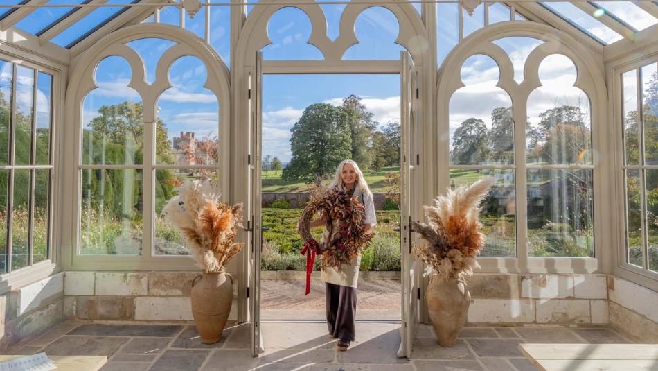 A person holding a dried flower wreath in the doorway of a sunny conservatory