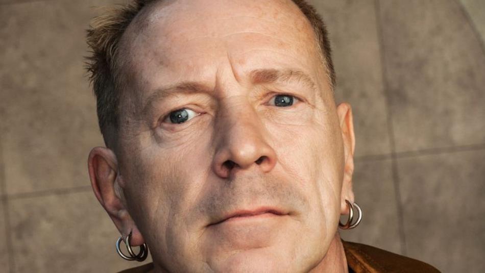 Close up of John Lydon looking quizzical with hoop earrings, with a beige background.