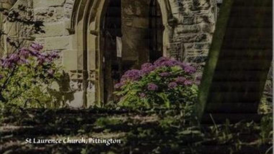 An arched stone church doorway, surrounded by purple flowers in the sunlight.