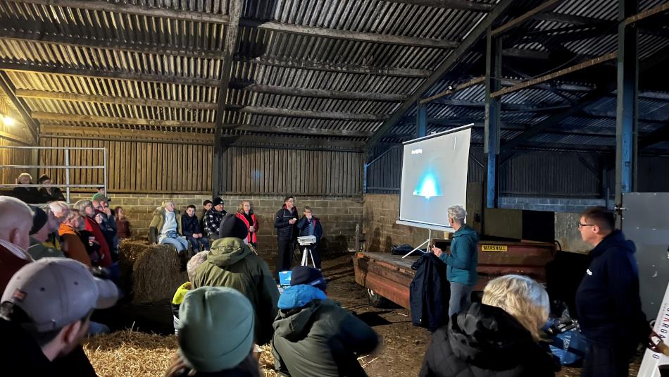 A group of people sit on hay bales inside a barn while a speaker gives an astronomy presentation beside a projected image.