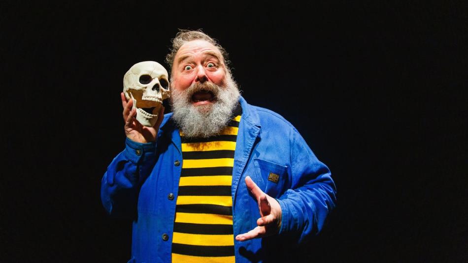 A bearded person, wearing a black and yellow striped top and blue jacket, screaming whilst holding a skull next to their face on a black background.